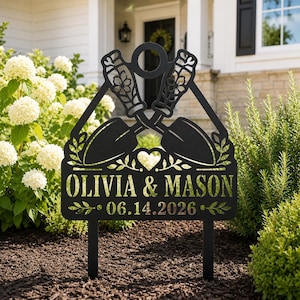 Könnte beinhalten: Ein schwarzer Gartenstecker aus Metall mit einem Scherenschnitt-Design aus zwei gekreuzten Handschaufeln, einem zentralen Herz und Blattmotiven. Der Text OLIVIA & MASON und das Datum 06.14.2026 sind in den unteren Teil des Schildes gelasert.