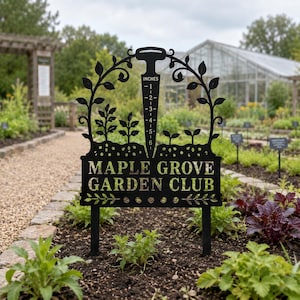 May include: A black metal garden sign featuring a central trowel shape with a ruler marked in inches from one to six. The sign displays the text MAPLE GROVE GARDEN CLUB, surrounded by decorative vine and leaf silhouettes on two metal stakes.