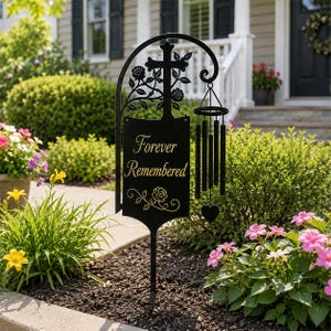 May include: A black metal garden stake featuring a cross and rose motif with a hanging wind chime. The central rectangular plaque displays the text "Forever Remembered" in gold lettering, accented by a decorative rose vine illustration at the base.
