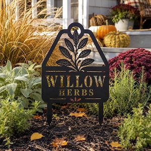Pode incluir: Uma estaca de jardim em metal preto com topo pontiagudo e recorte circular, apresentando um design cortado a laser de um ramo de planta com folhas acima do texto "WILLOW HERBS" em fonte serifada, com duas pontas na base para fixação.