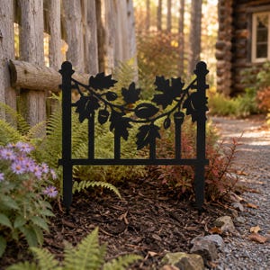 May include: A black metal garden border fence panel featuring a decorative cutout design of oak leaves and acorns. The rectangular panel has two vertical posts with rounded finials at the top and pointed stakes at the bottom for ground insertion.
