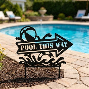 Op de afbeelding: Een zwart metalen tuinbord in de vorm van een pijl die naar rechts wijst. Het bord bevat de tekst POOL THIS WAY in sjabloonletters, met een strandbalafbeelding boven de tekst en een reddingsboei in watergolven onder de tekst.