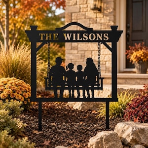 May include: Black metal family sign featuring a swing with a family silhouette. The sign reads "THE WILSONS" in a cutout font. The sign is designed to be placed in a garden or yard, with two stakes at the bottom.