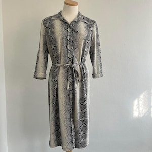May include: A long-sleeved dress with a snakeskin pattern in shades of gray and cream. The dress features a collared neckline, button-down front, and a matching belt at the waist. The sleeves are three-quarter length.