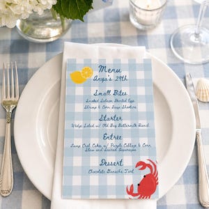 May include: A menu card with a blue and white gingham pattern, featuring the text "Menu Anya's 29th." The menu lists small bites, a starter, an entrée, and dessert. A lemon and a red crab are illustrated on the card. The card is on a white plate.