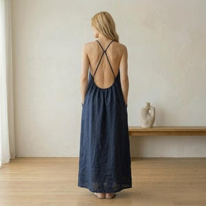 Könnte beinhalten: Ein marineblaues Maxikleid mit tiefem Rückenausschnitt und gekreuzten Trägern. Das Kleid hat eine lockere Passform mit Taschen und einem bodenlangen Saum. Das Model steht in einem hellen Raum mit neutralem Hintergrund.