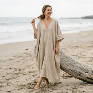 Peut inclure: Une robe caftan en lin beige avec un col en V et des manches larges. La robe est longue et portée sur une plage de sable. Une grosse bûche repose sur le sable près de la robe.