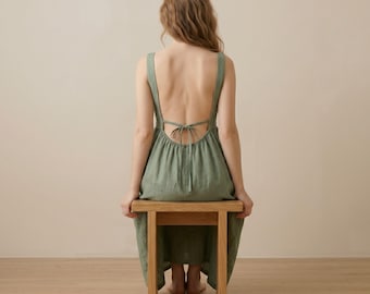 Puede incluir: Un vestido de lino verde salvia con espalda baja y detalle de lazo. El vestido tiene tirantes anchos y una falda fluida. La persona está sentada en un taburete de madera clara. El fondo es beige neutro.