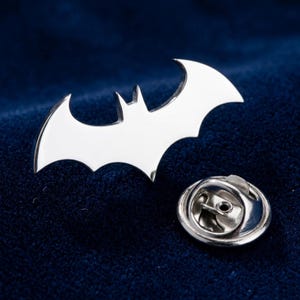 May include: A silver-colored Batman logo pin with a butterfly clutch. The pin features the iconic bat symbol, a stylized silhouette of a bat with outstretched wings. The pin is set against a dark blue background.