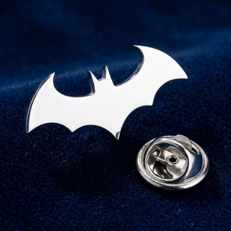 May include: A silver-colored Batman logo pin with a butterfly clutch. The pin features the iconic bat symbol, a stylized silhouette of a bat with outstretched wings. The pin is set against a dark blue background.