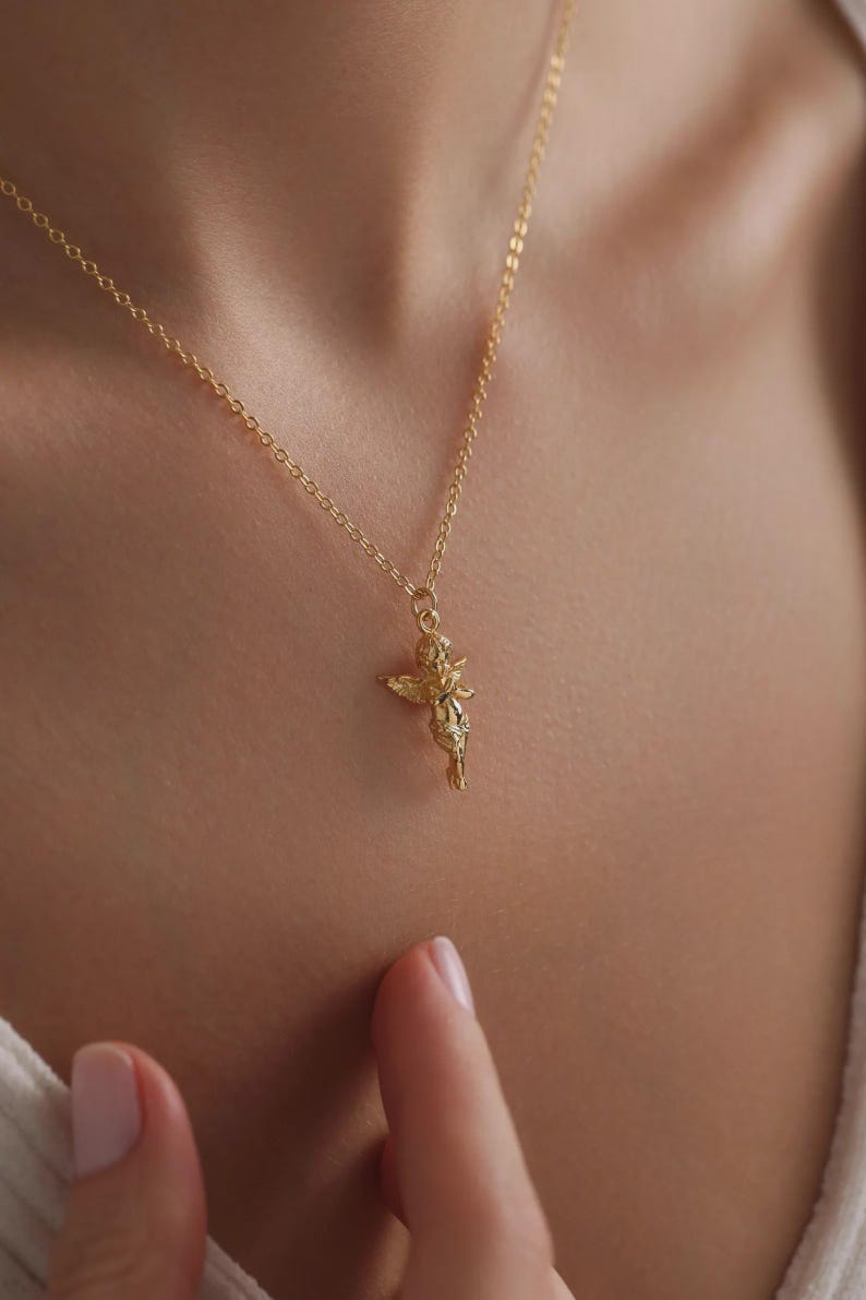 May include: A gold-coloured necklace with a small cherub pendant. The cherub has outstretched wings and hangs from a delicate chain. The necklace is worn against a skin tone background.