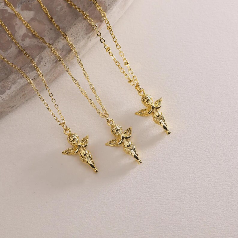 May include: Three gold necklaces, each with a cherub pendant. The necklaces have fine chains and detailed cherub charms with wings. The jewellery is displayed on a light-coloured surface.