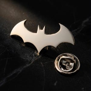 May include: A silver-colored metal pin in the shape of a bat, with a pin back. The bat emblem is a stylized design with pointed wings and a central body. The pin is likely for clothing or accessories.