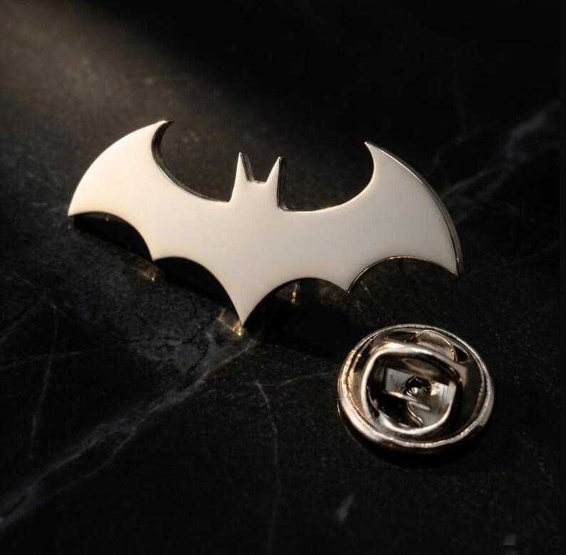 May include: A silver-colored metal pin in the shape of a bat, with a pin back. The bat emblem is a stylized design with pointed wings and a central body. The pin is likely for clothing or accessories.