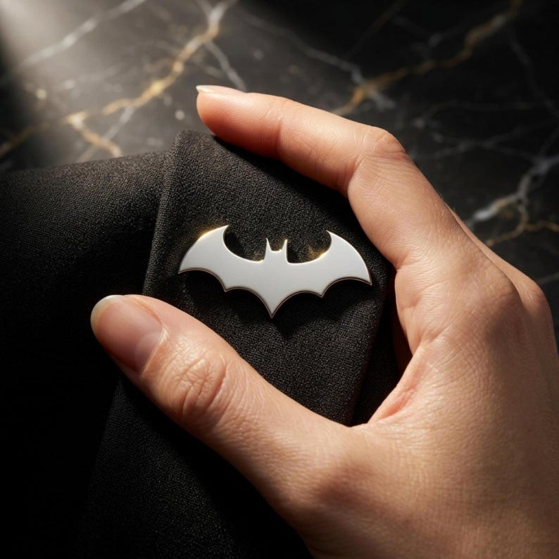 May include: A silver-colored Batman logo pin. The pin is in the shape of a bat with outstretched wings. The pin is attached to a black fabric. The pin is held by a hand.