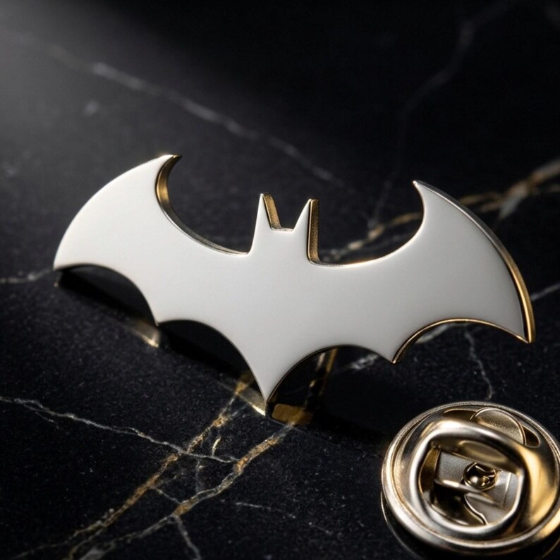 May include: A silver-colored Batman logo pin with gold-colored trim. The pin is in the shape of a bat with outstretched wings. The pin is on a black marble surface, with a gold-colored pin back.