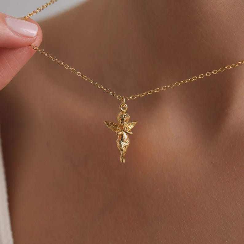 May include: A gold necklace with a cherub pendant. The pendant is a detailed miniature figure with wings, hanging from a fine chain. The necklace is displayed against a plain backdrop, emphasizing the gold colour and design.