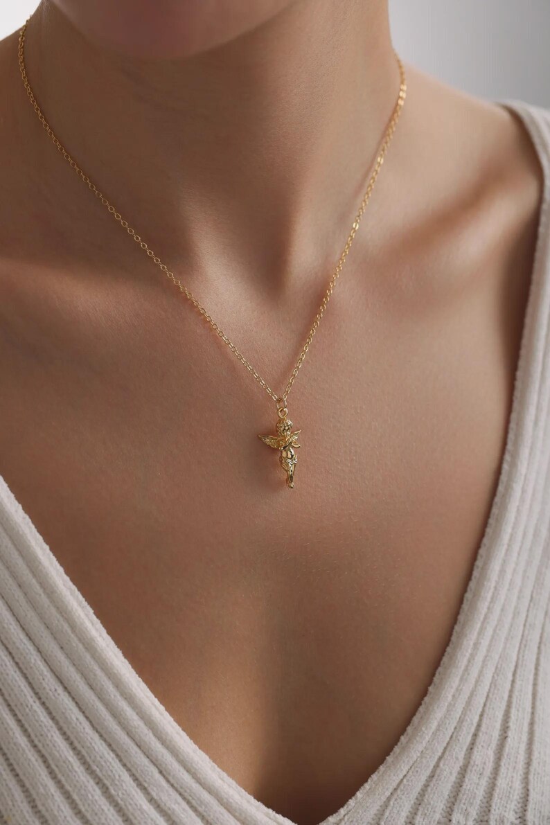 May include: A gold-coloured necklace with a small cherub pendant. The cherub has detailed wings and hangs from a delicate chain. The necklace is shown against a neutral background, emphasising the jewellery's design.