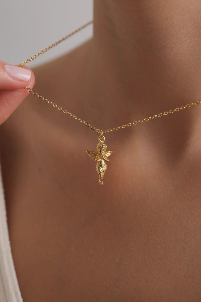 May include: A gold-coloured necklace with a cherub pendant. The cherub has detailed wings and clasped hands. The necklace chain is delicate. The jewellery is displayed against a neutral background. The pendant is a small, detailed sculpture.