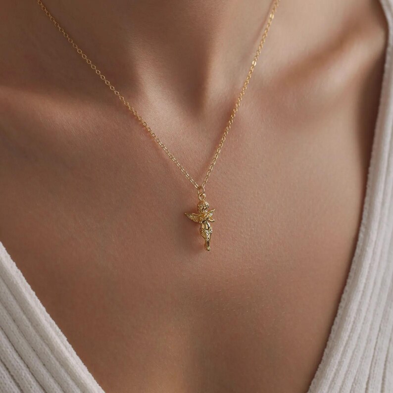 May include: A gold-coloured necklace with a small cherub pendant. The cherub has detailed wings and hangs from a delicate chain. The necklace is worn against a white top.