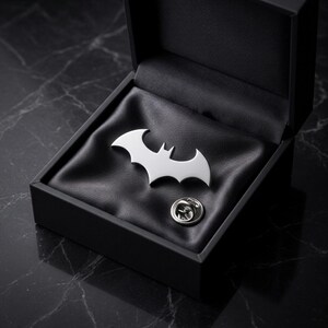 May include: A silver-colored Batman logo pin displayed in an open black gift box with a satin lining. The pin is in the shape of a bat with outstretched wings. A silver pin back is also visible.