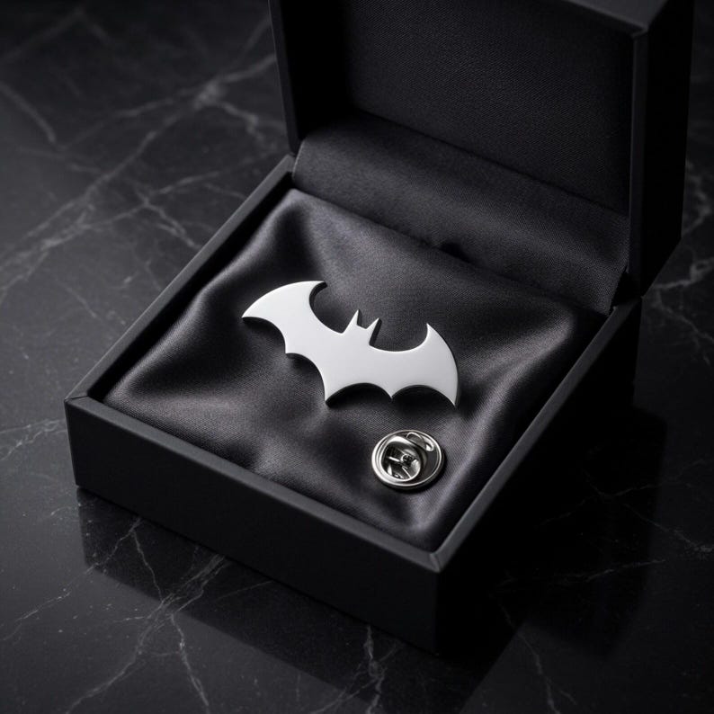 May include: A silver-colored Batman logo pin displayed in an open black gift box with a satin lining. The pin is in the shape of a bat with outstretched wings. A silver pin back is also visible.