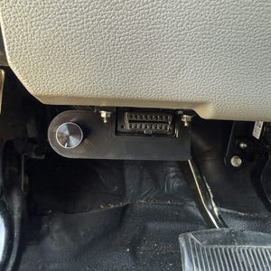 May include: A black OBD2 port mount with a silver knob is installed under a vehicle's dashboard. The mount is secured with bolts and is positioned near the gas pedal. The dashboard is a light gray color.