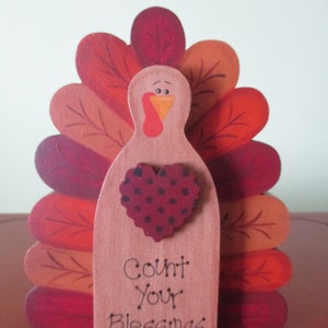 Op de afbeelding: Houten kalkoenfiguur met een rode, oranje en bruine geschilderde staartveren. De kalkoen heeft een hart met zwarte stippen en de woorden "Count Your Blessings" op de voorkant.