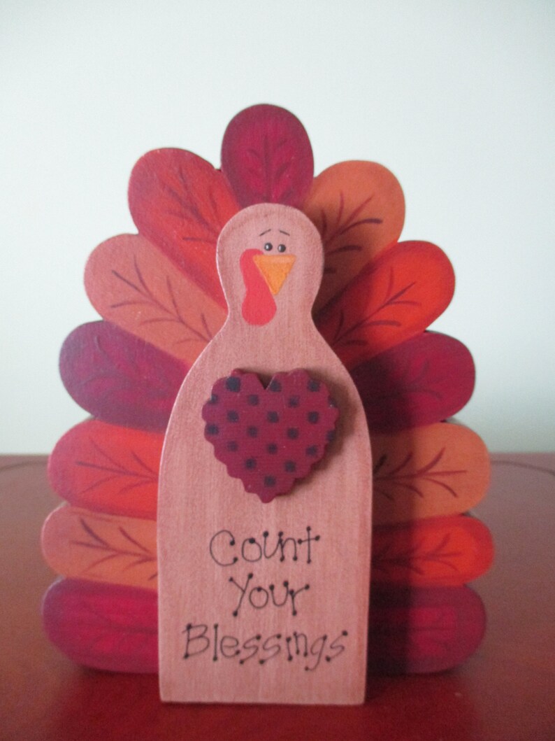 Op de afbeelding: Houten kalkoenfiguur met een rode, oranje en bruine geschilderde staartveren. De kalkoen heeft een hart met zwarte stippen en de woorden "Count Your Blessings" op de voorkant.