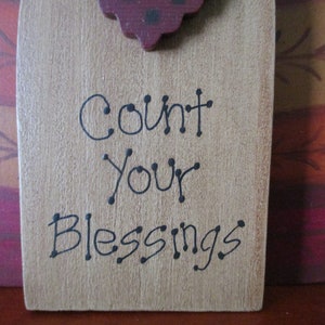 Op de afbeelding: Een houten bord met een bruine achtergrond en zwarte tekst die "Count Your Blessings" luidt. Het bord is versierd met een rood en zwart kalkoenhoofd aan de bovenkant.
