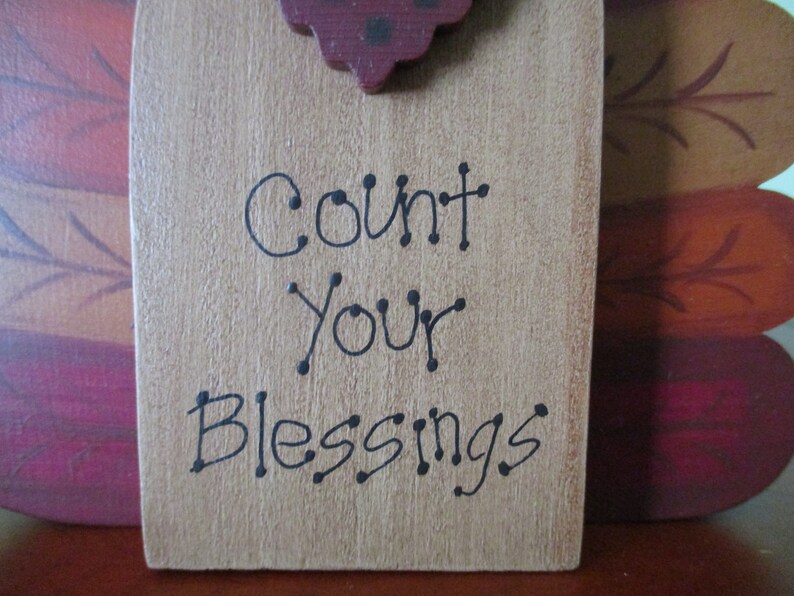 Op de afbeelding: Een houten bord met een bruine achtergrond en zwarte tekst die "Count Your Blessings" luidt. Het bord is versierd met een rood en zwart kalkoenhoofd aan de bovenkant.