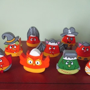 May include: A collection of ten hand-painted wooden candy corn figurines, each depicting a different Halloween character. The characters include a witch, a pirate, a vampire, a bat, an owl, a Frankenstein, a mummy, and a cat. The figurines are all painted in bright colors and have whimsical expressions.