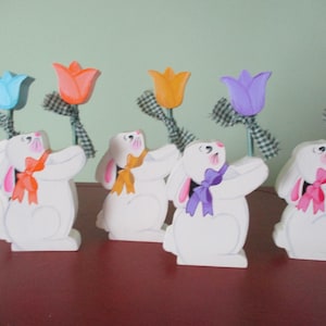 May include: Five white wooden bunny figurines holding colorful painted tulips. The bunnies are painted white with pink, blue, purple, orange, and pink bows. The tulips are painted blue, orange, purple, pink, and orange.