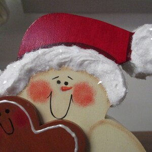 Puede incluir: Una figurita de mu&ntilde;eco de nieve de madera que lleva un gorro de Pap&aacute; Noel rojo con ribete blanco, sosteniendo una galleta de jengibre en forma de coraz&oacute;n. El mu&ntilde;eco de nieve tiene una cara pintada con mejillas sonrosadas y una nariz negra.