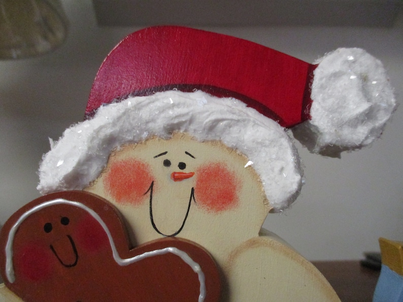 Puede incluir: Una figurita de mu&ntilde;eco de nieve de madera que lleva un gorro de Pap&aacute; Noel rojo con ribete blanco, sosteniendo una galleta de jengibre en forma de coraz&oacute;n. El mu&ntilde;eco de nieve tiene una cara pintada con mejillas sonrosadas y una nariz negra.
