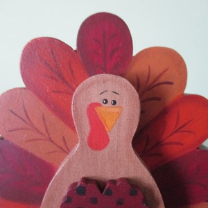 Op de afbeelding: Houten kalkoenfiguur met een rood, oranje en bruin geschilderde staartveren. De kalkoen heeft een bruin lichaam, een rode keelzak en een oranje snavel.