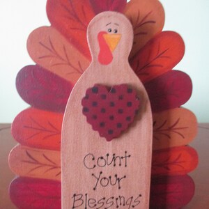 Op de afbeelding: Een houten kalkoenfiguur met een rood, oranje en bruin geschilderd ontwerp. De kalkoen heeft een hartvormig ontwerp met zwarte stippen en de tekst "Count Your Blessings".