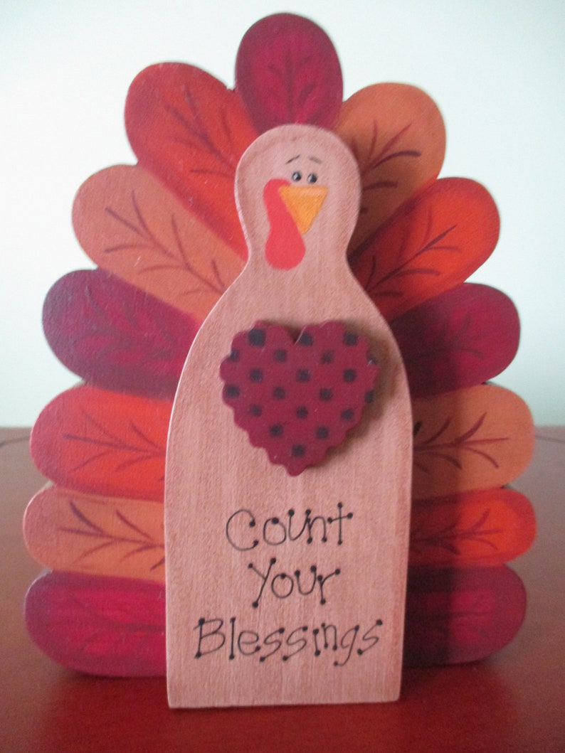 Op de afbeelding: Een houten kalkoenfiguur met een rood, oranje en bruin geschilderd ontwerp. De kalkoen heeft een hartvormig ontwerp met zwarte stippen en de tekst "Count Your Blessings".