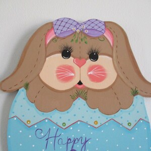 May include: A painted wooden Easter bunny with brown fur, a purple bow, and a blue and white polka dot egg-shaped body. The bunny has a pink nose and whiskers, and the egg says "Happy".