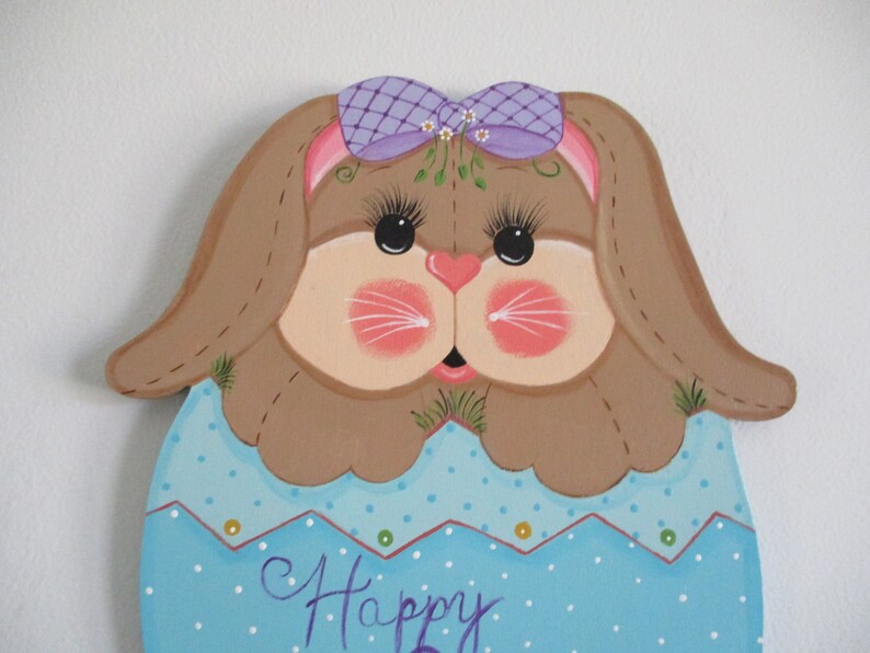 May include: A painted wooden Easter bunny with brown fur, a purple bow, and a blue and white polka dot egg-shaped body. The bunny has a pink nose and whiskers, and the egg says "Happy".