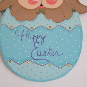 May include: A wooden Easter bunny decoration painted in brown, blue, and white. The bunny is holding an egg with white polka dots and the words "Happy Easter" written in purple.
