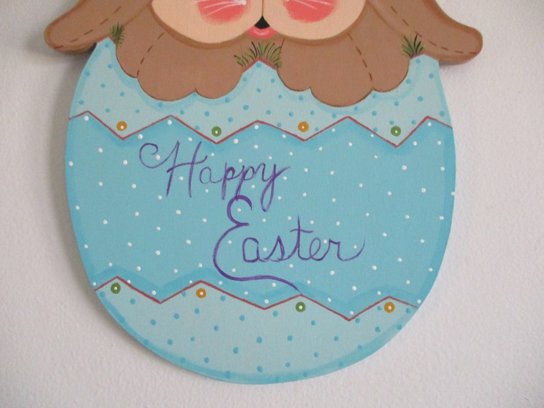 May include: A wooden Easter bunny decoration painted in brown, blue, and white. The bunny is holding an egg with white polka dots and the words "Happy Easter" written in purple.