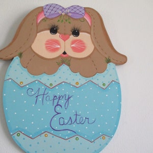 May include: A wooden Easter bunny cutout painted in brown, pink, and purple. The bunny is wearing a purple bow and has a blue egg with white polka dots and the text "Happy Easter" painted on it.