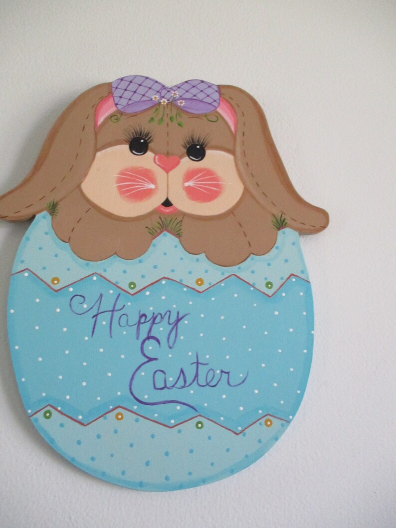 May include: A wooden Easter bunny cutout painted in brown, pink, and purple. The bunny is wearing a purple bow and has a blue egg with white polka dots and the text "Happy Easter" painted on it.