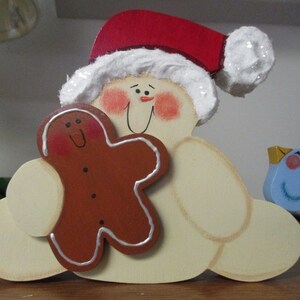 Puede incluir: Una figura de madera de mu&ntilde;eco de nieve con un gorro de Pap&aacute; Noel rojo, sosteniendo un hombre de jengibre y un p&aacute;jaro azul. El mu&ntilde;eco de nieve tiene una barba blanca y mejillas sonrosadas.