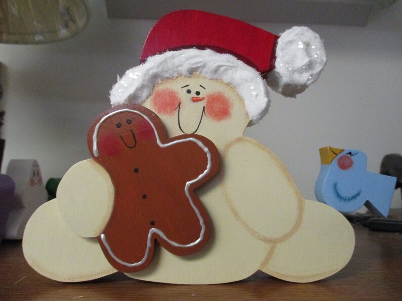 Puede incluir: Una figura de madera de mu&ntilde;eco de nieve con un gorro de Pap&aacute; Noel rojo, sosteniendo un hombre de jengibre y un p&aacute;jaro azul. El mu&ntilde;eco de nieve tiene una barba blanca y mejillas sonrosadas.
