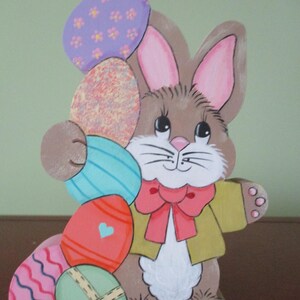May include: Wooden Easter bunny holding colorful painted eggs. The bunny is brown with pink ears and a red bow tie. The eggs are decorated with various patterns and colors, including purple flowers, blue stripes, and a heart.