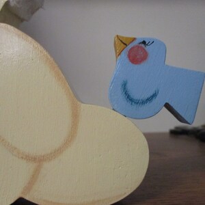 Puede incluir: Una figurita de p&aacute;jaro de madera con un cuerpo azul claro, un pico amarillo y ojos negros. El p&aacute;jaro est&aacute; posado sobre un objeto de madera m&aacute;s grande de color crema.