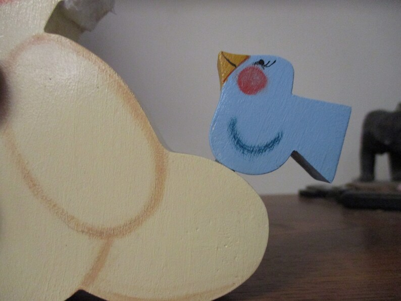 Puede incluir: Una figurita de p&aacute;jaro de madera con un cuerpo azul claro, un pico amarillo y ojos negros. El p&aacute;jaro est&aacute; posado sobre un objeto de madera m&aacute;s grande de color crema.