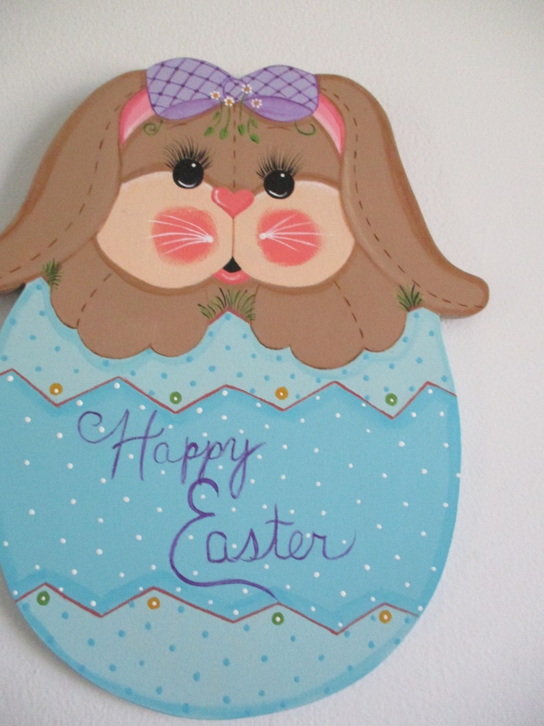 May include: A painted wooden Easter bunny with a purple bow and a blue egg with white polka dots. The egg says "Happy Easter" in purple paint.